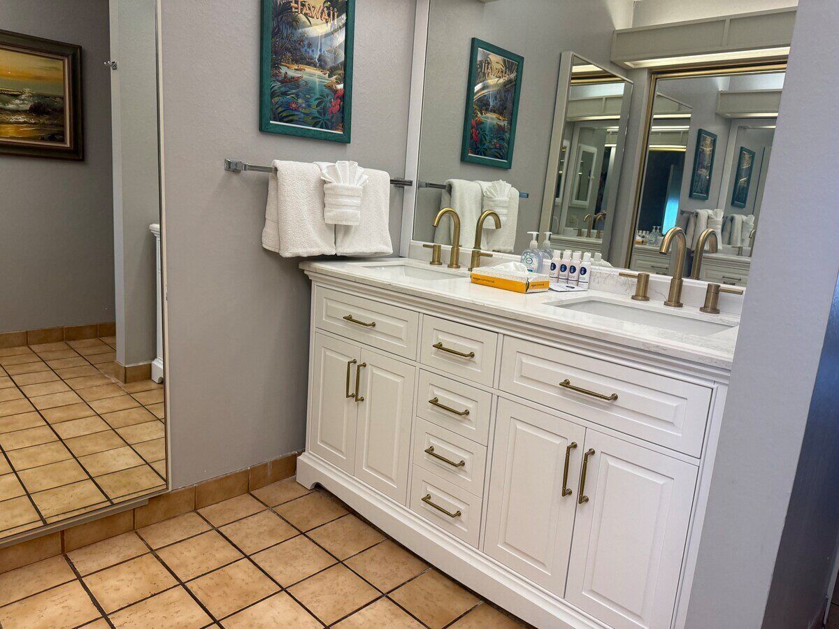 Une salle de bain avec un meuble-lavabo blanc, des robinets dorés et un grand miroir. Deux tableaux encadrés sont accrochés au mur.