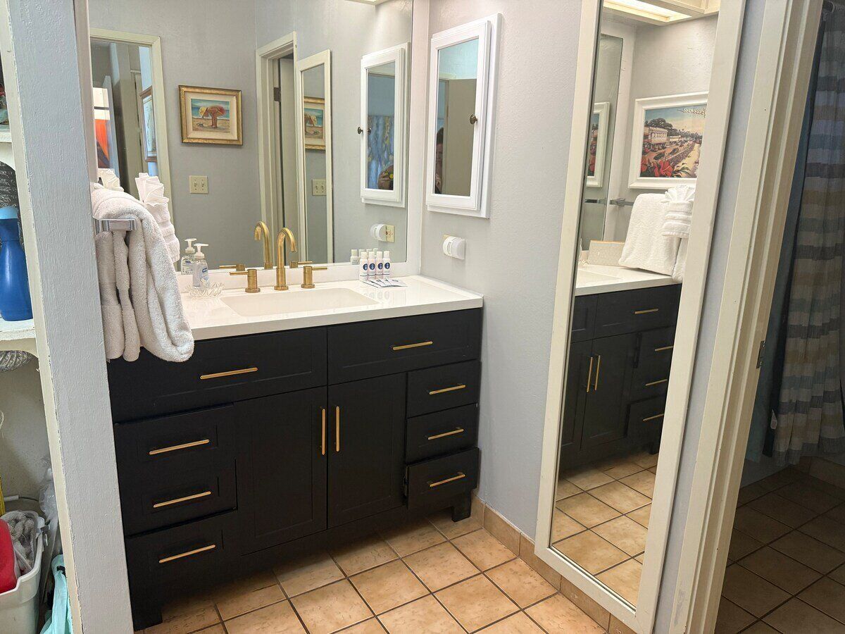 A bathroom with a dark vanity, white sink, and two mirrors. There are towels hanging on the wall and a shower curtain visible in the background.