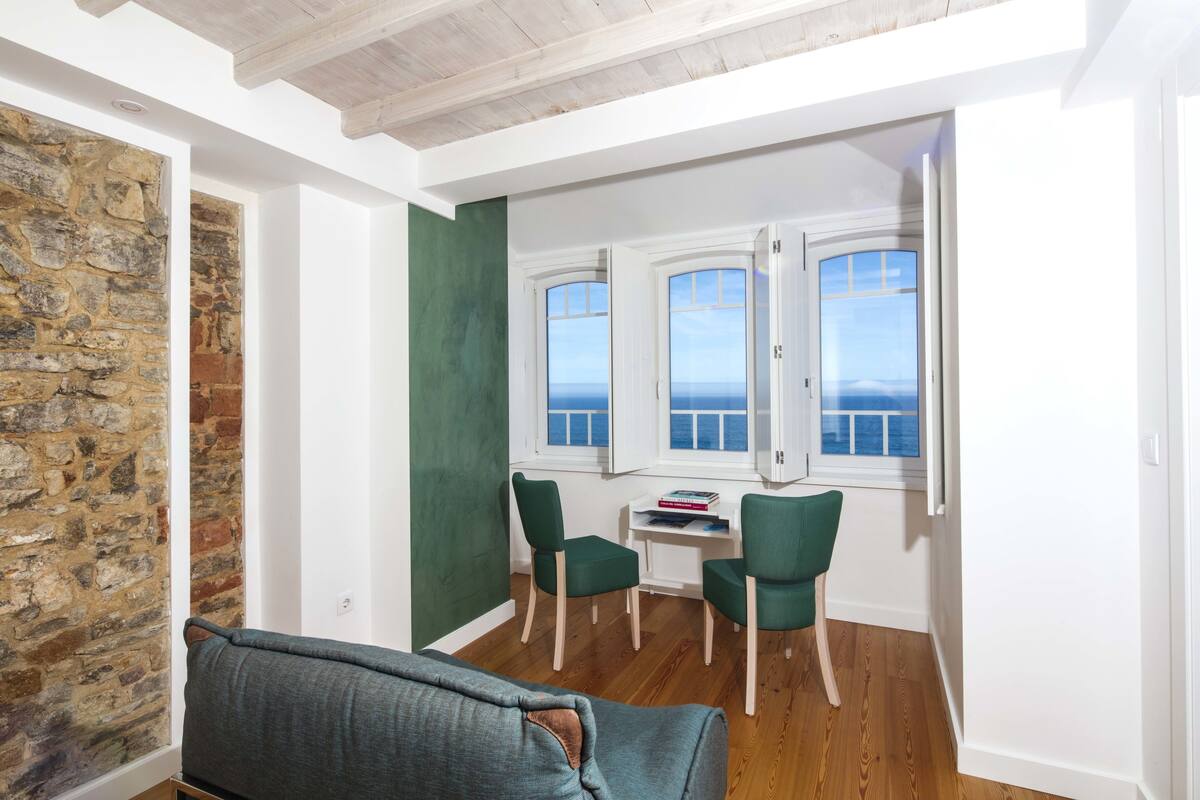 A cozy room with a stone wall, a green wall, and a wooden ceiling. There are two green chairs and a table with books.