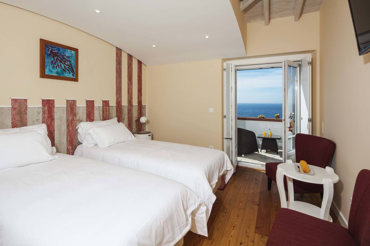 A hotel room with two beds, a small table with a chair, a view of the sea, and a balcony.