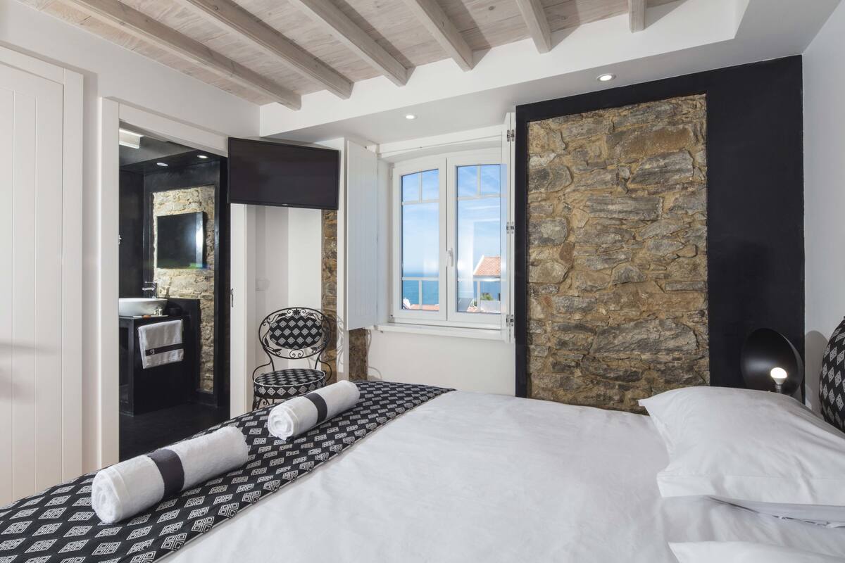A bedroom with a bed, a stone accent wall, a view of the sea, a bathroom with a sink, and a chair.