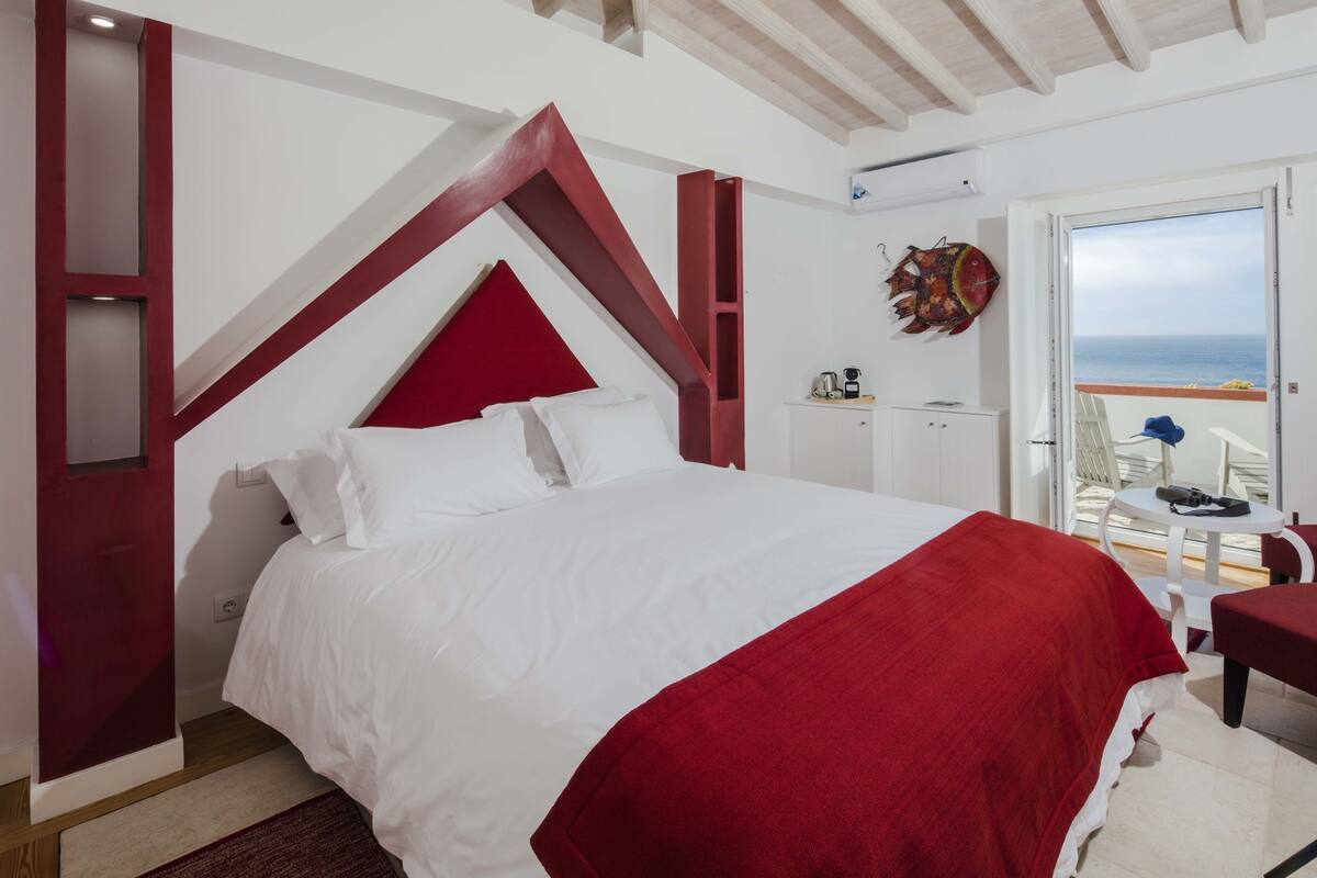 A bedroom with a large bed, a red accent wall, a view of the sea through an open door, and a small table with a chair.