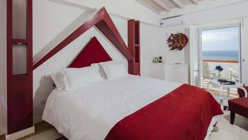 A bedroom with a large bed, a red accent wall, a view of the sea through an open door, and a small table with a chair.