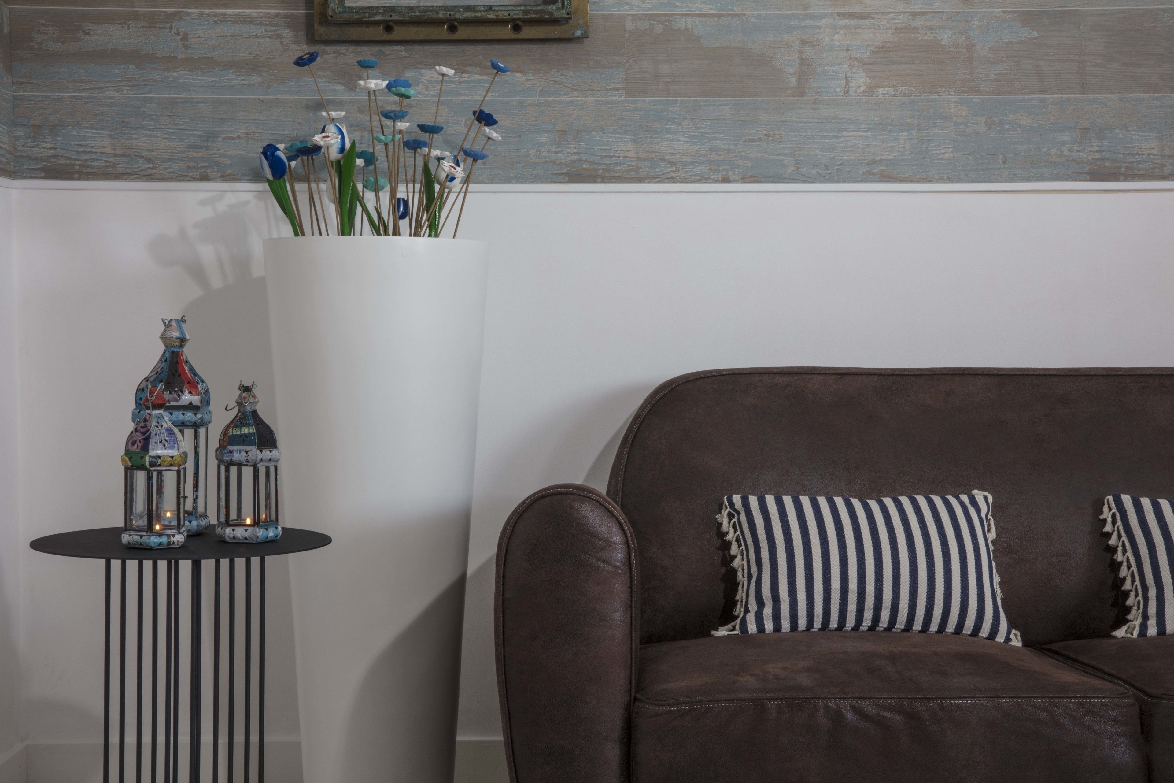 Een bruine leren bank met gestreepte kussens, een witte vaas met blauwe bloemen en twee decoratieve vogeltafels op een zwart bijzettafeltje.