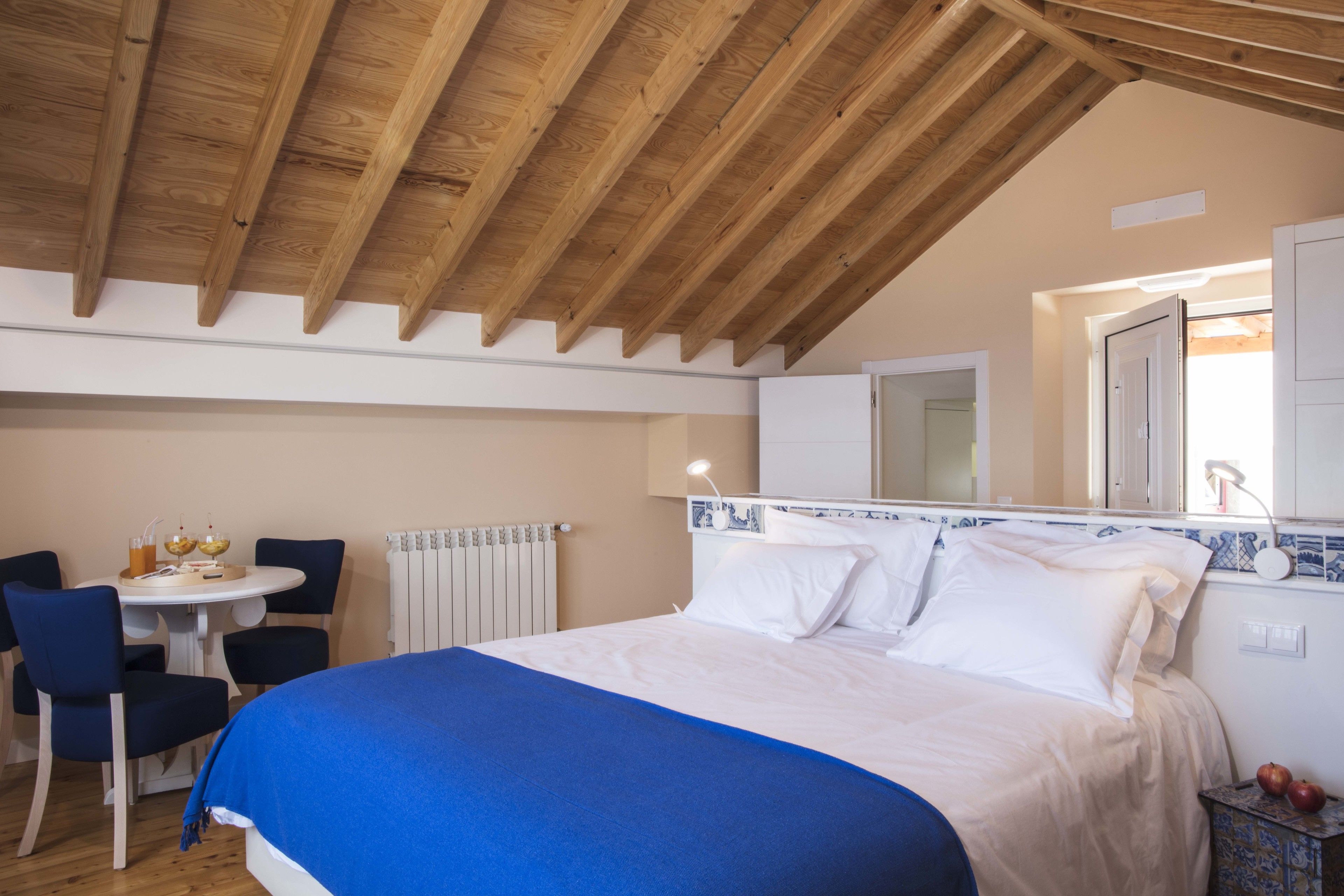 Quarto com teto de madeira, cama com roupa de cama branca, uma cadeira azul, uma mesinha com bebidas e um radiador.