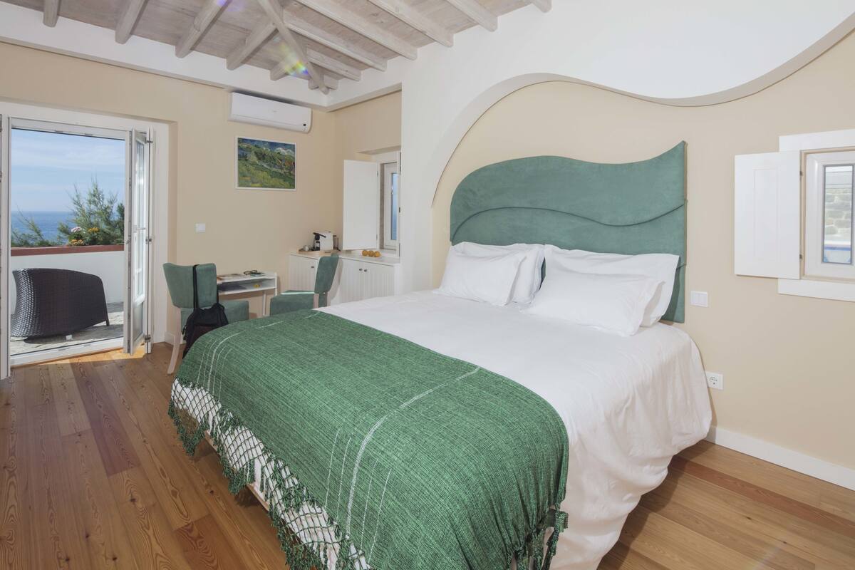 A bedroom with a large bed, a desk, and a chair. There is a balcony with a view of the sea.