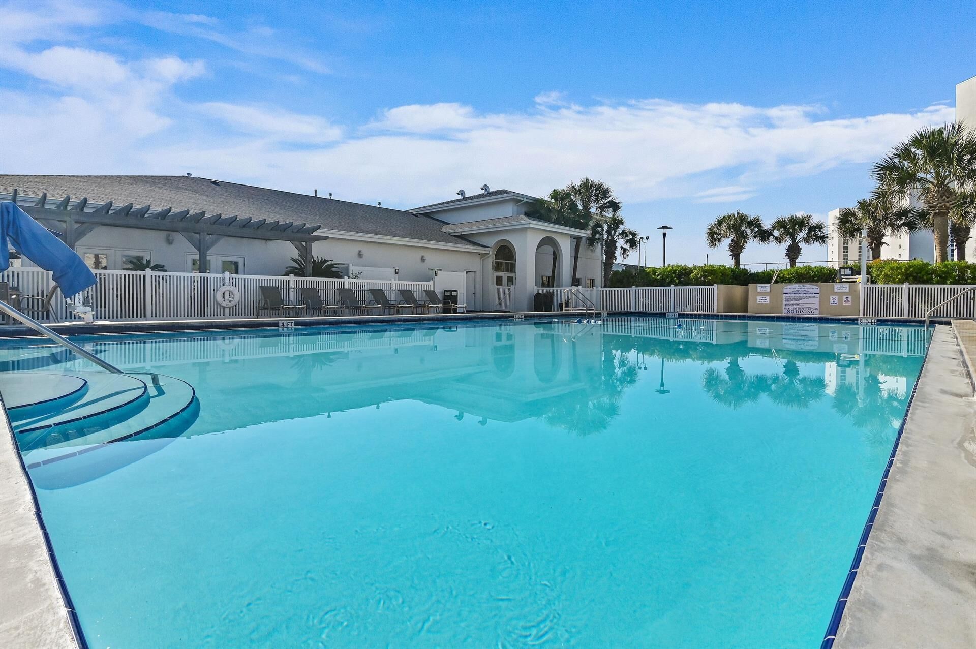 A swimming pool with a clear blue water, surrounded by a white fence, and a building with a grey roof in the background.