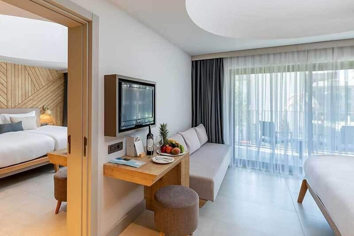 Una habitación de hotel moderna con cama, escritorio con televisor, zona de estar y un ventanal con cortinas.
