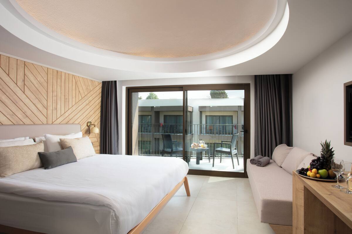 Una habitación de hotel moderna con una cama grande, un sofá, una mesa de comedor con frutas y copas de vino, y un balcón con vistas al edificio.
