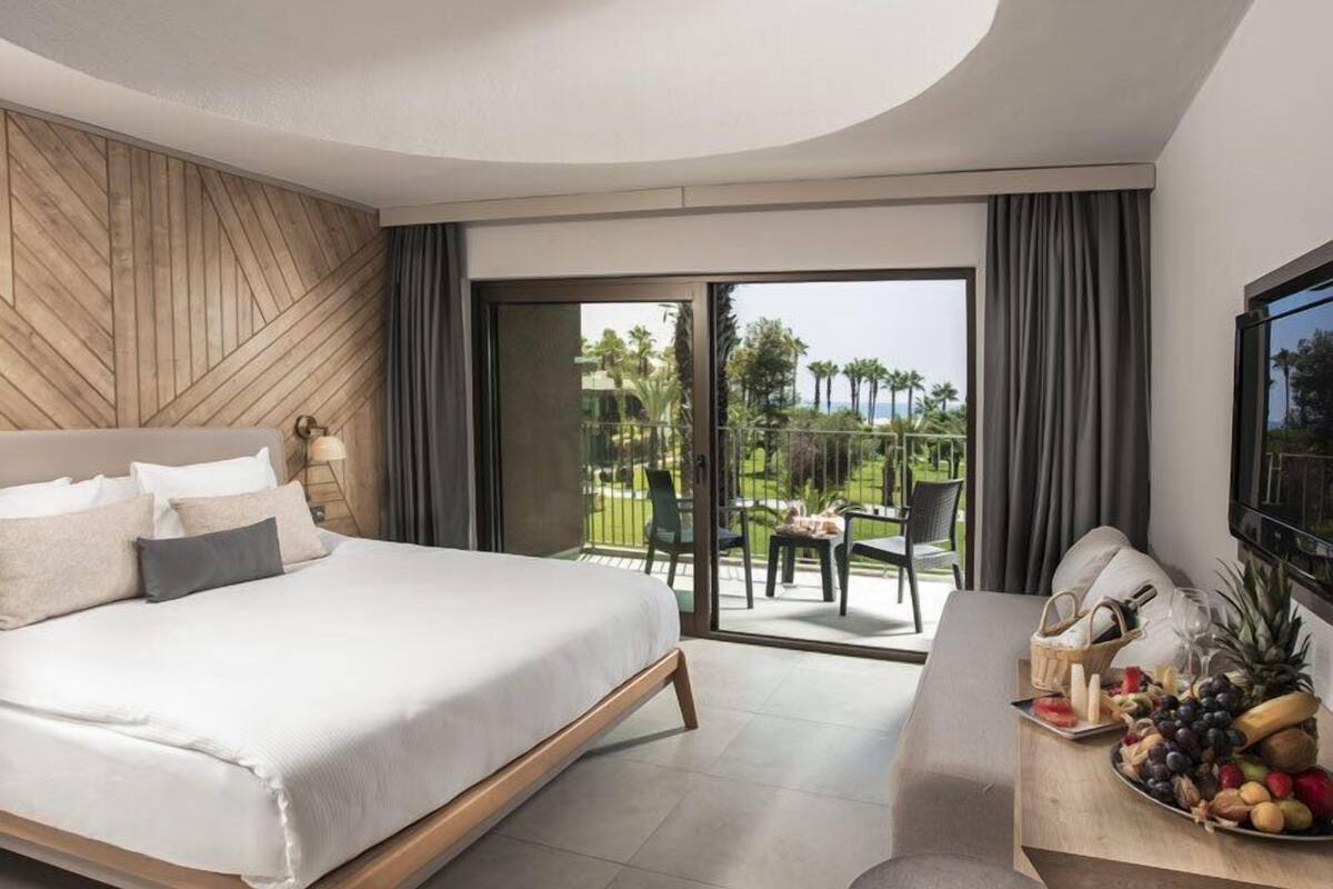 Una habitación de hotel con cama, una zona de comedor con cesta de frutas y un balcón con vistas a palmeras y al océano.