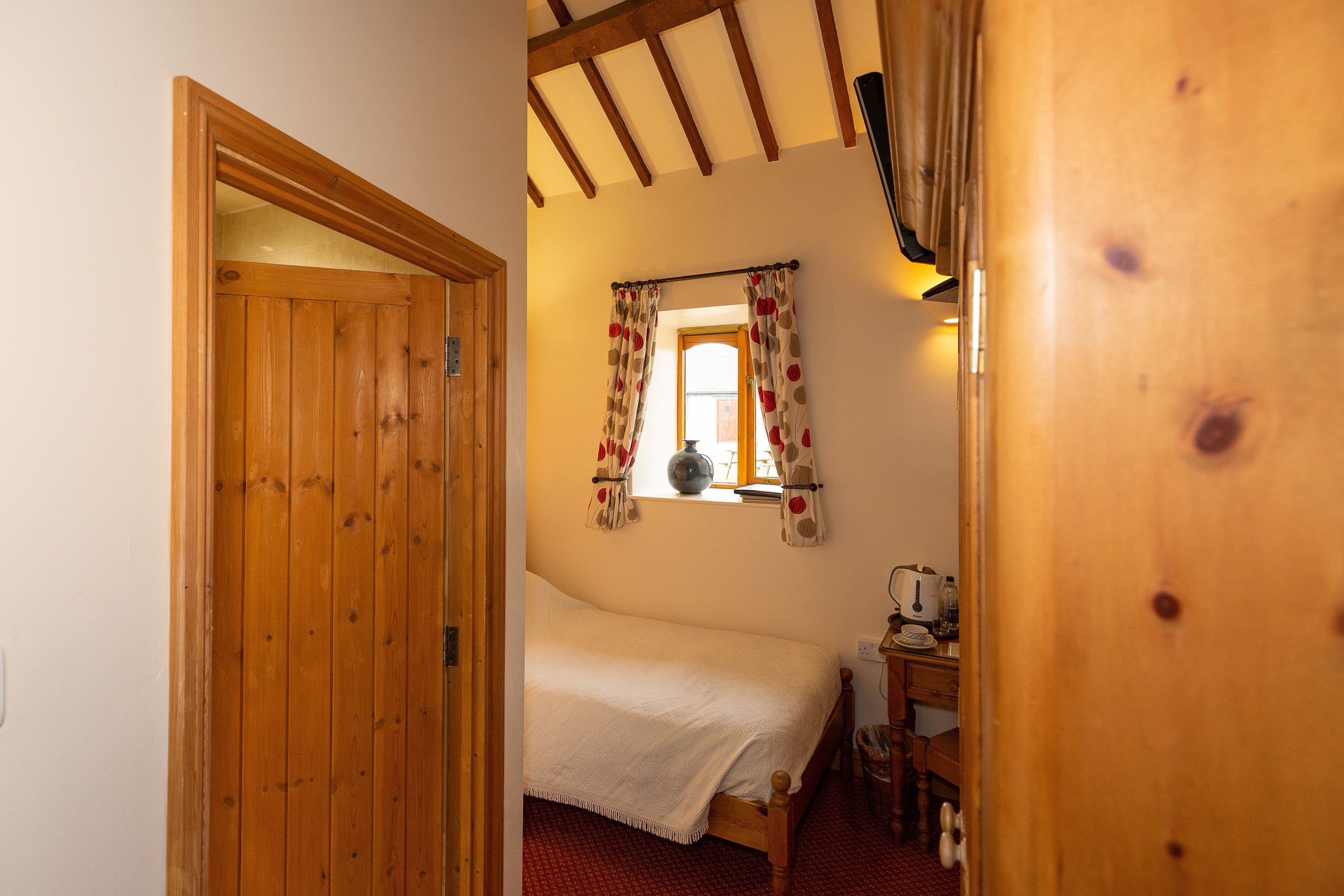 Une porte en bois donne sur une chambre à coucher qui contient un lit, une fenêtre avec des rideaux et un bureau avec un téléphone.
