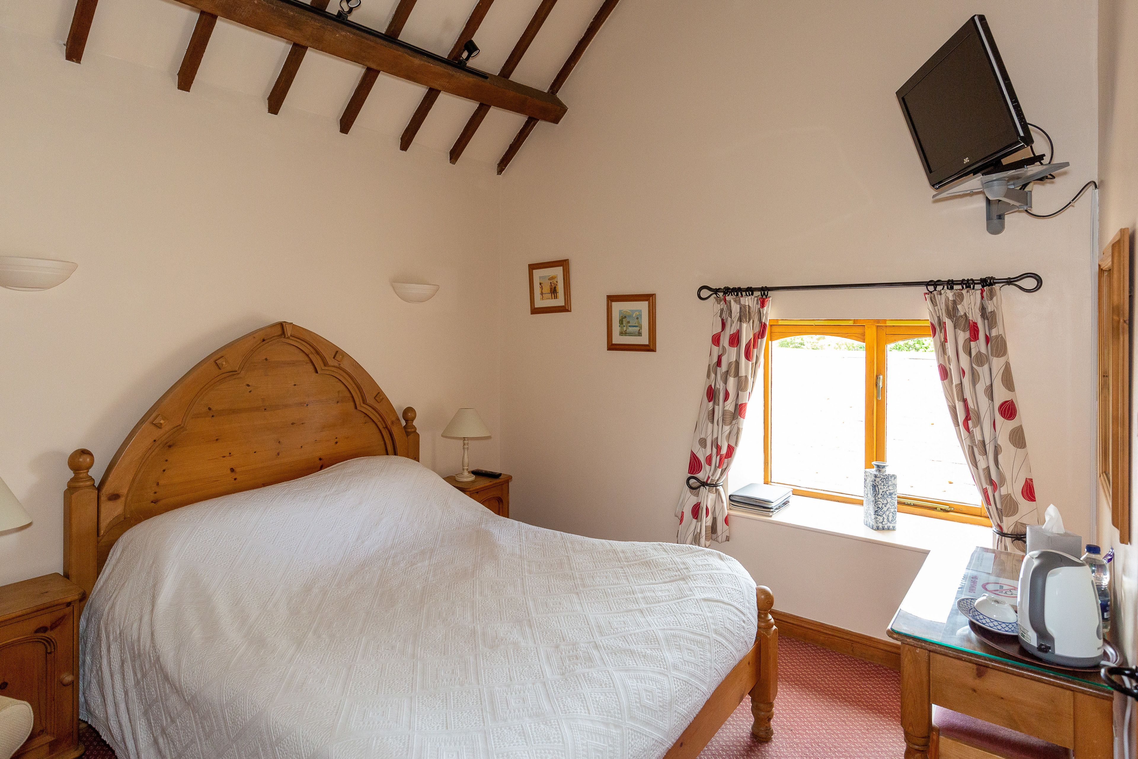 A bedroom with a wooden bed, a desk with a kettle, a television mounted on the wall, and a window with floral curtains.