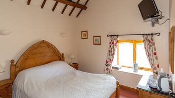 A bedroom with a wooden bed, a desk with a kettle, a television mounted on the wall, and a window with floral curtains.