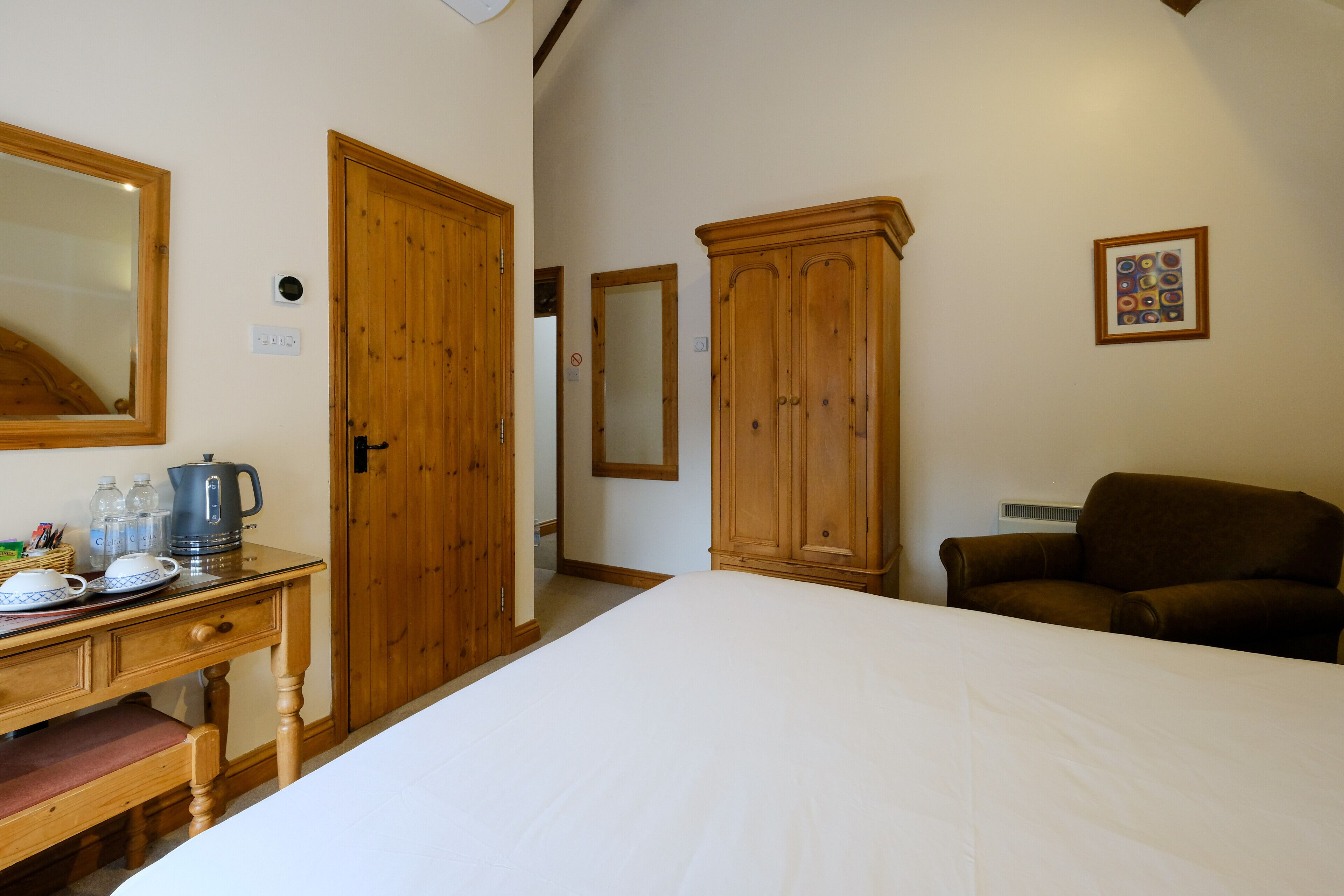 Quarto com cama, guarda-roupa de madeira, secretária com chaleira e poltrona marrom.