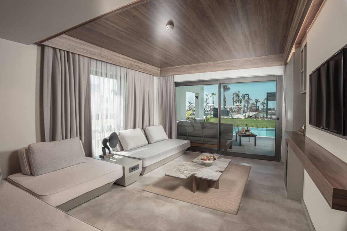 A modern living room with a sofa, coffee table, and a view of a pool and palm trees through large glass doors.