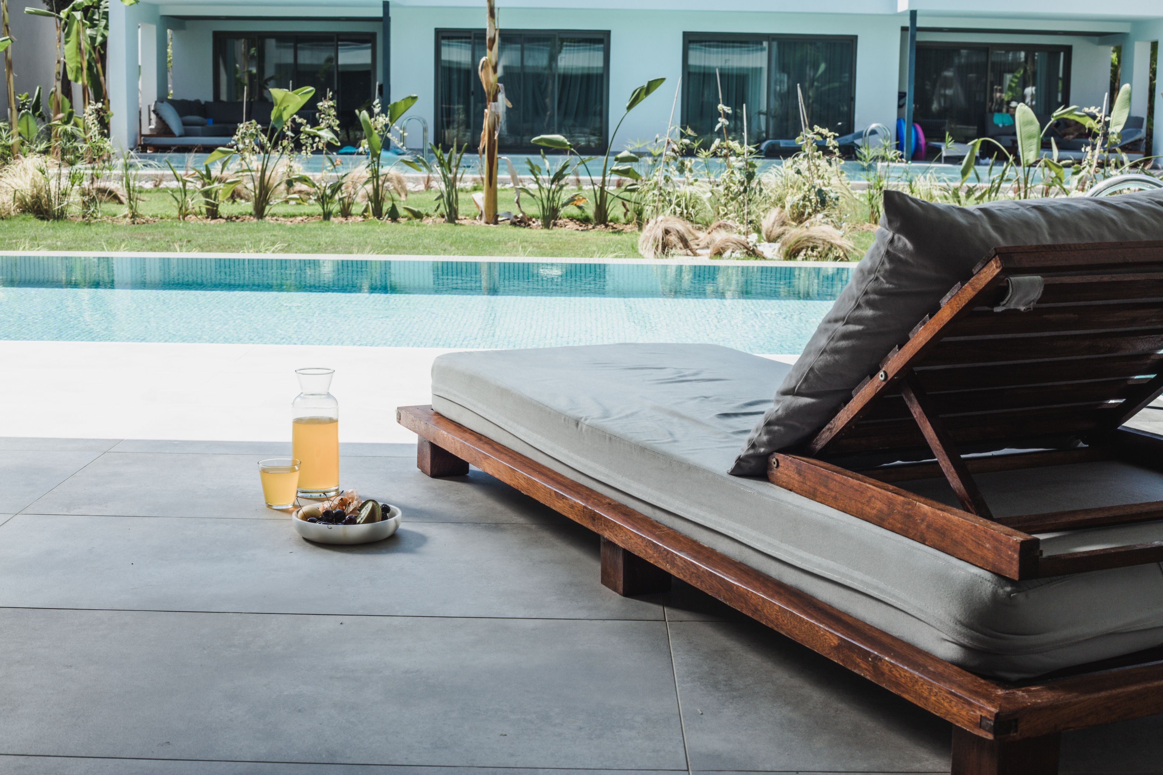 A poolside lounge area with a wooden bench and cushions, a glass of beverage, and a bowl of snacks.