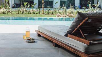 A poolside lounge area with a wooden bench and cushions, a glass of beverage, and a bowl of snacks.