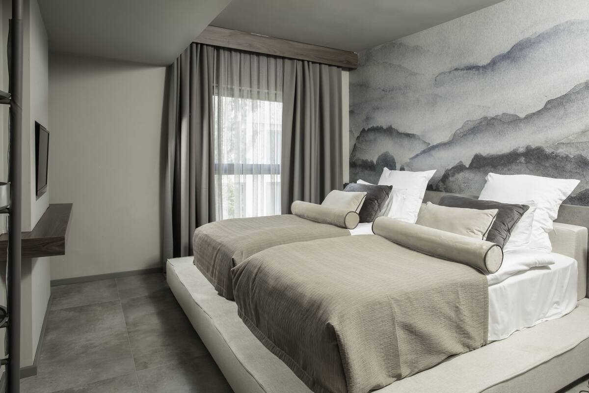 A modern bedroom with two large beds, a large window with curtains, a wall-mounted TV, and a painting of mountains.