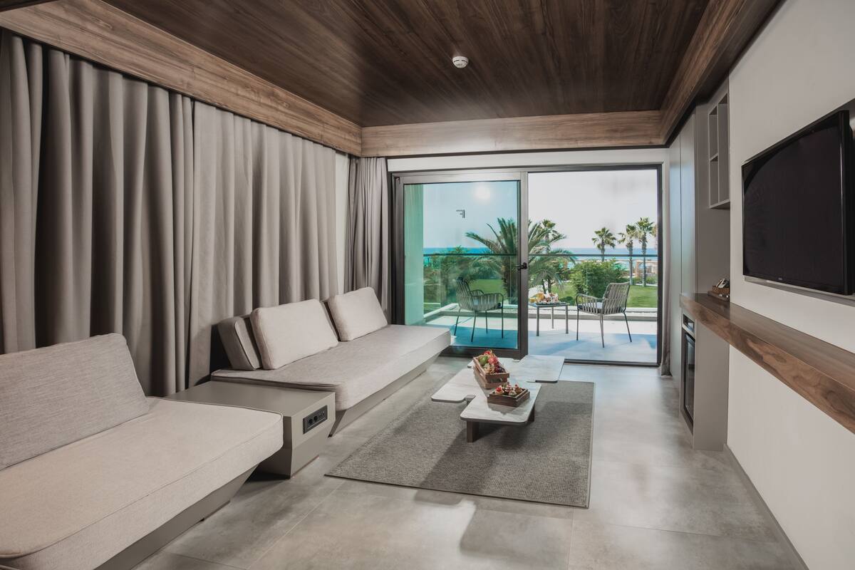 A modern living room with a sofa, a coffee table, and a TV. There is a sliding glass door leading to a balcony with a view of palm trees and the ocean.