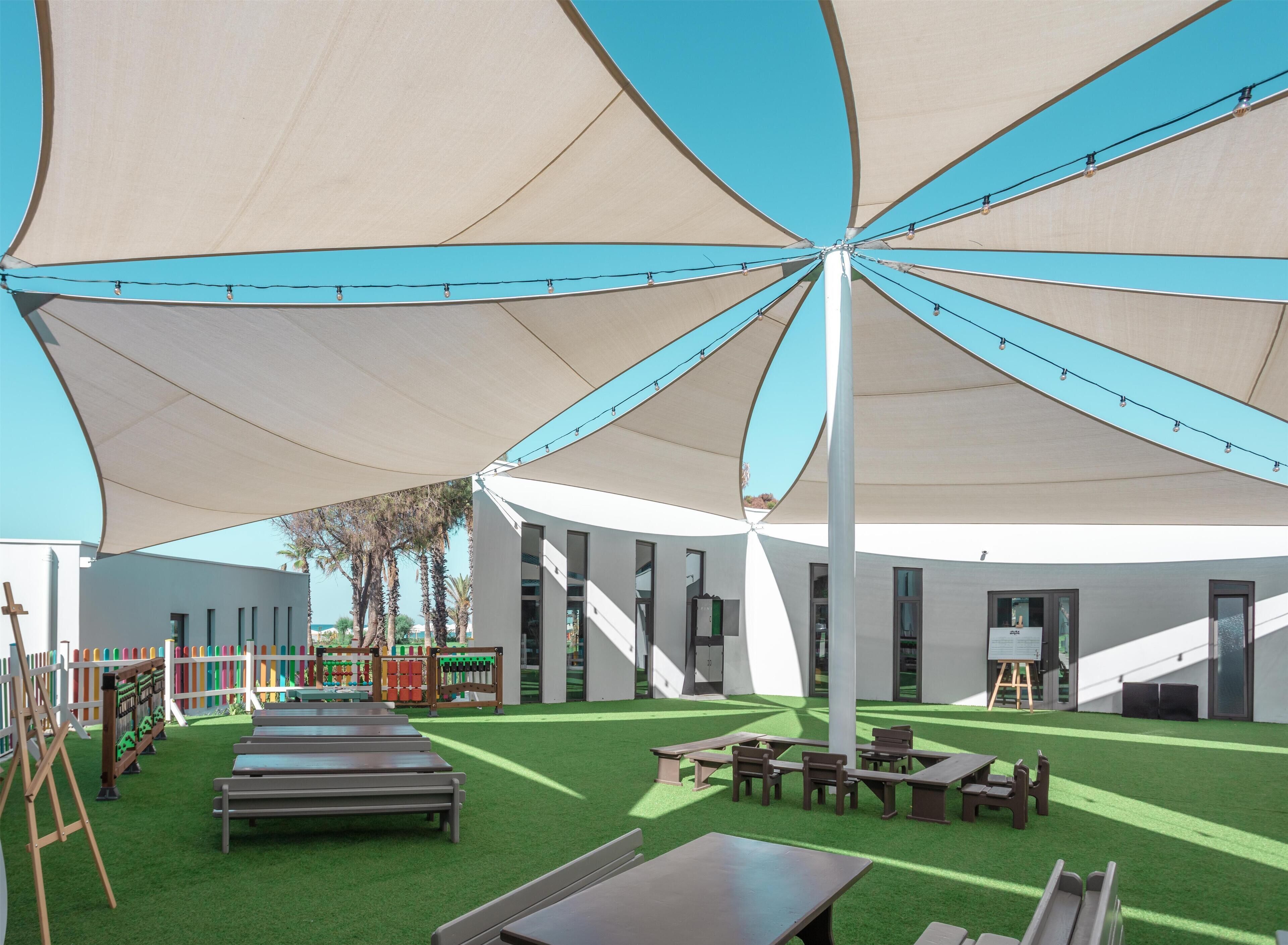 Un espacio al aire libre con una gran estructura tipo dosel, césped artificial y varias mesas y bancos de madera.