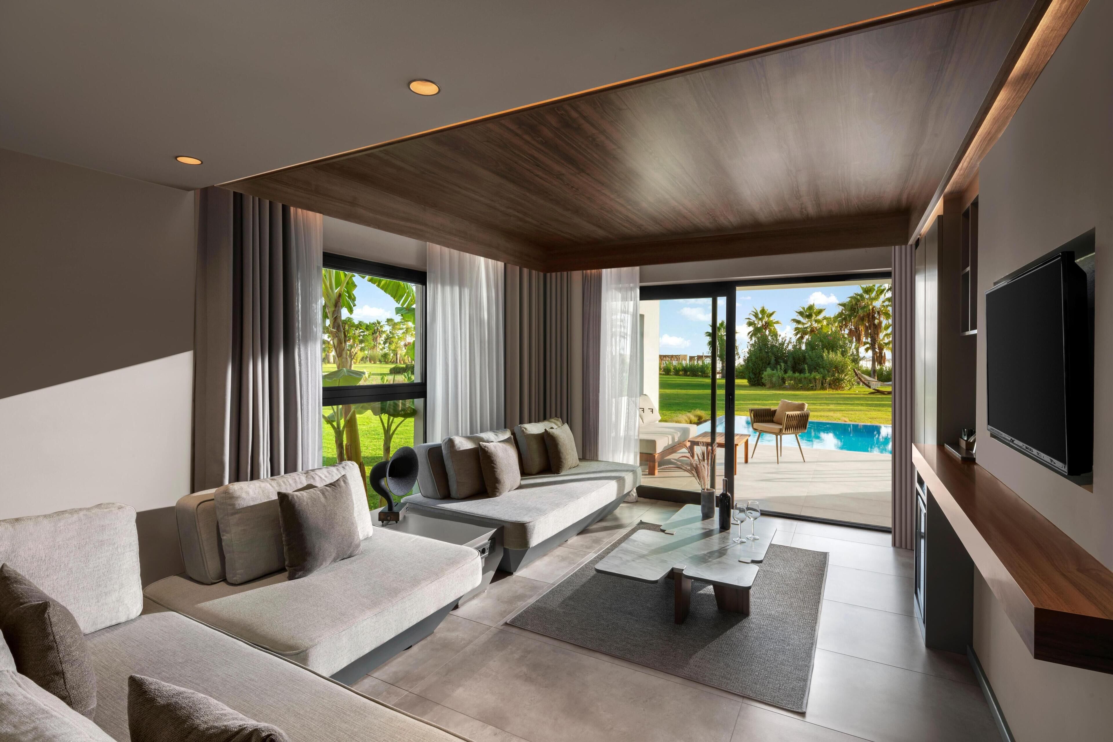 Uma sala de estar moderna com um sofá, mesa de centro e televisão. Há uma porta de vidro deslizante que dá para uma área de piscina com mobiliário exterior.