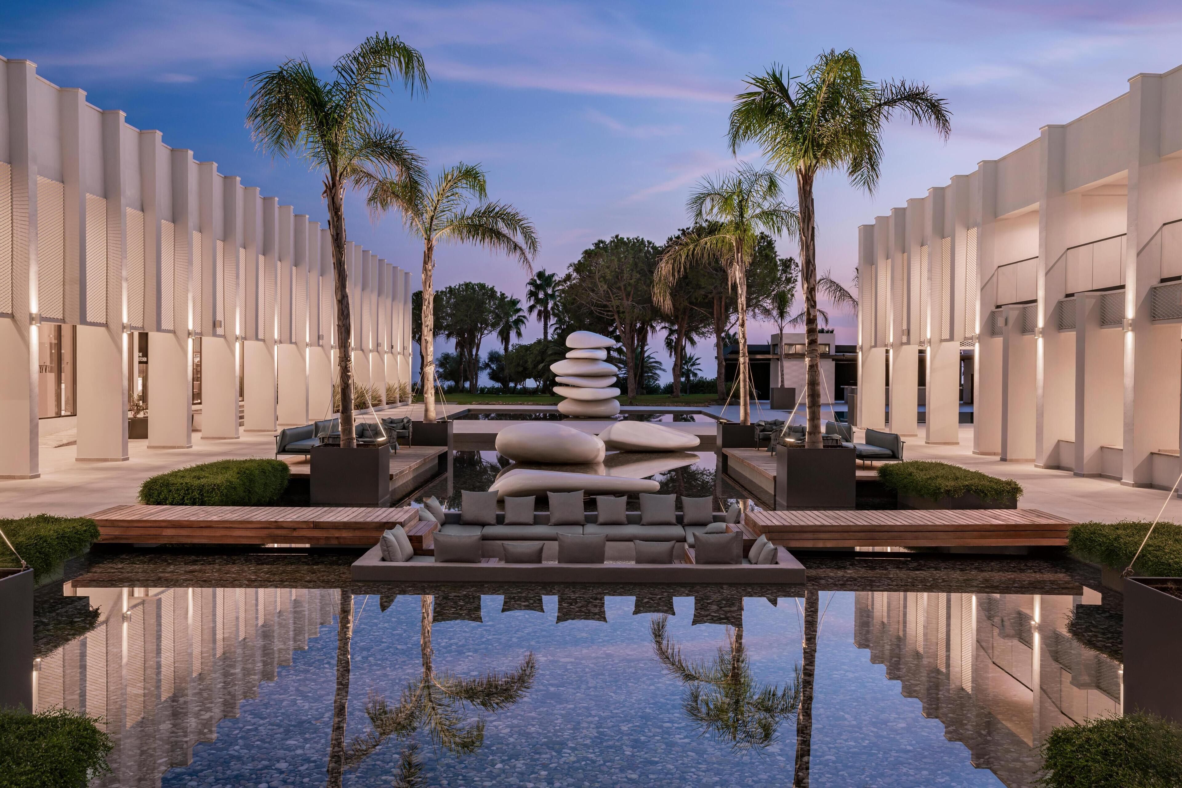 Un hotel moderno con una piscina reflectante, palmeras y una escultura rocosa singular.