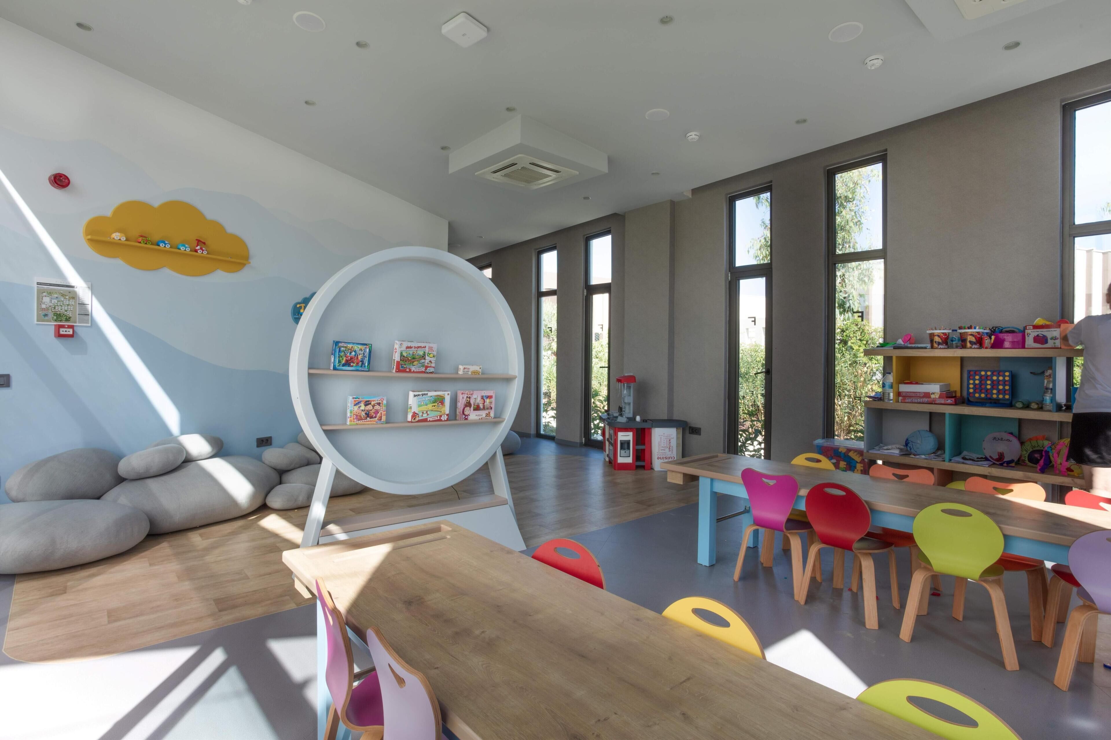 Una habitación amplia con ventanas grandes, una estantería circular y sillas de colores.