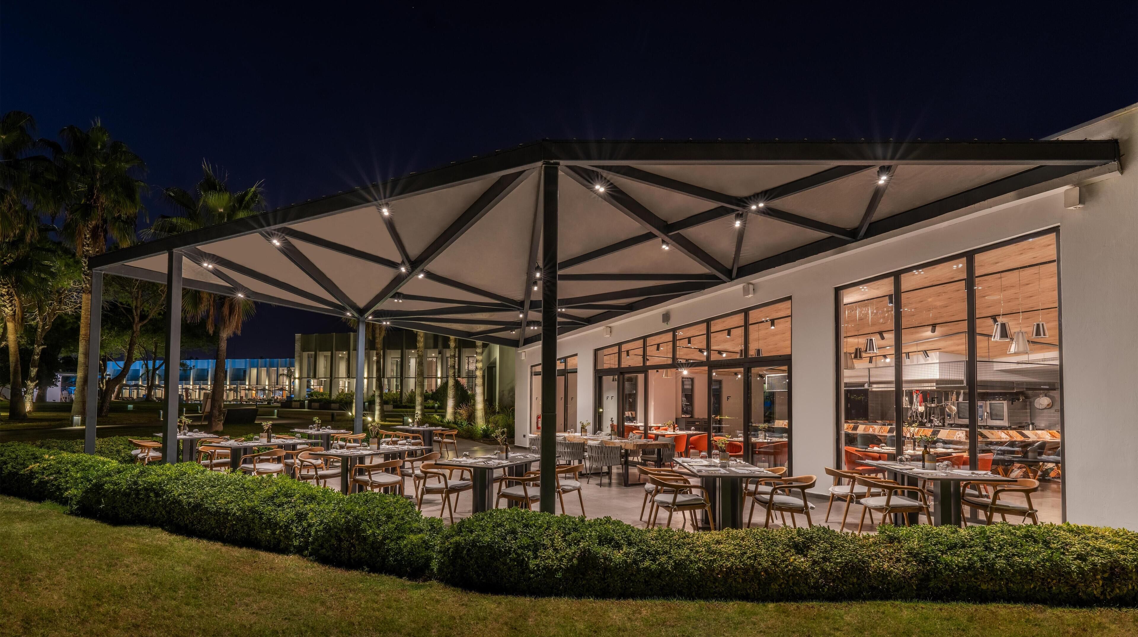 An outdoor dining area with tables and chairs, a covered seating area, and a view of the kitchen through large windows.