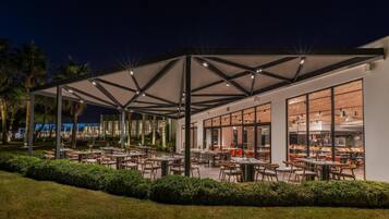 An outdoor dining area with tables and chairs, a covered seating area, and a view of the kitchen through large windows.