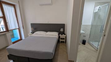 A bedroom with a bed, bedside tables, a lamp, an air conditioning unit, a window with curtains, and a shower area.