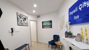 A hotel reception area with a digital clock, a reception desk, a blue chair, a bench, and artwork on the walls. - Napoli City Inn (Naples)