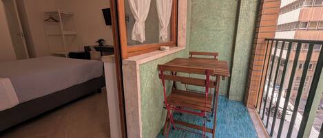 A bedroom with a bed, a small table, a window with curtains, and a balcony with a railing.