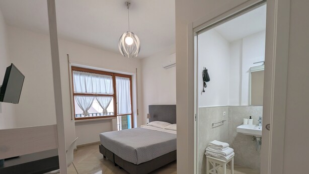 A bedroom with a bed, a bedside table, a hanging light fixture, a window with curtains, and a bathroom with a sink and towel rack. - Napoli City Inn (Naples)