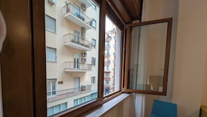 A room with a window overlooking a multi-story building, a blue chair, and a wooden floor. - Napoli City Inn (Naples)