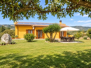 A yellow villa with a tiled roof, a patio with a table and chairs, and a view of the surrounding landscape.