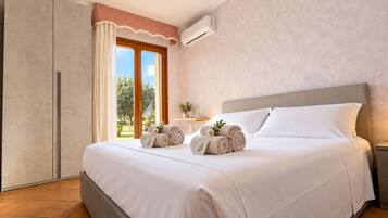 A hotel room with a bed, a nightstand, an air conditioning unit, a window with curtains, and a view of greenery outside.