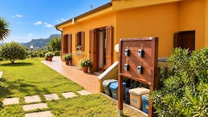 A yellow building with a paved walkway, wooden shutters, and potted plants.