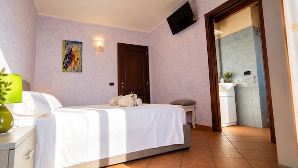 A hotel room with a bed, a bedside table, a lamp, a painting, a door, and a bathroom visible through an open door.