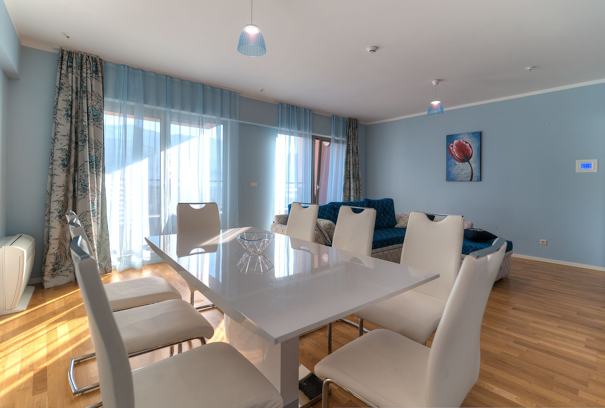 Een moderne woonkamer met een glazen eettafel, witte stoelen, een bank en een schilderij aan de muur.