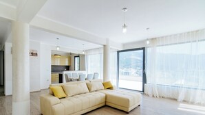 A modern living room with a beige sectional sofa, a dining area with white chairs, and large windows with sheer curtains. - Harmonia Colours (Budva)