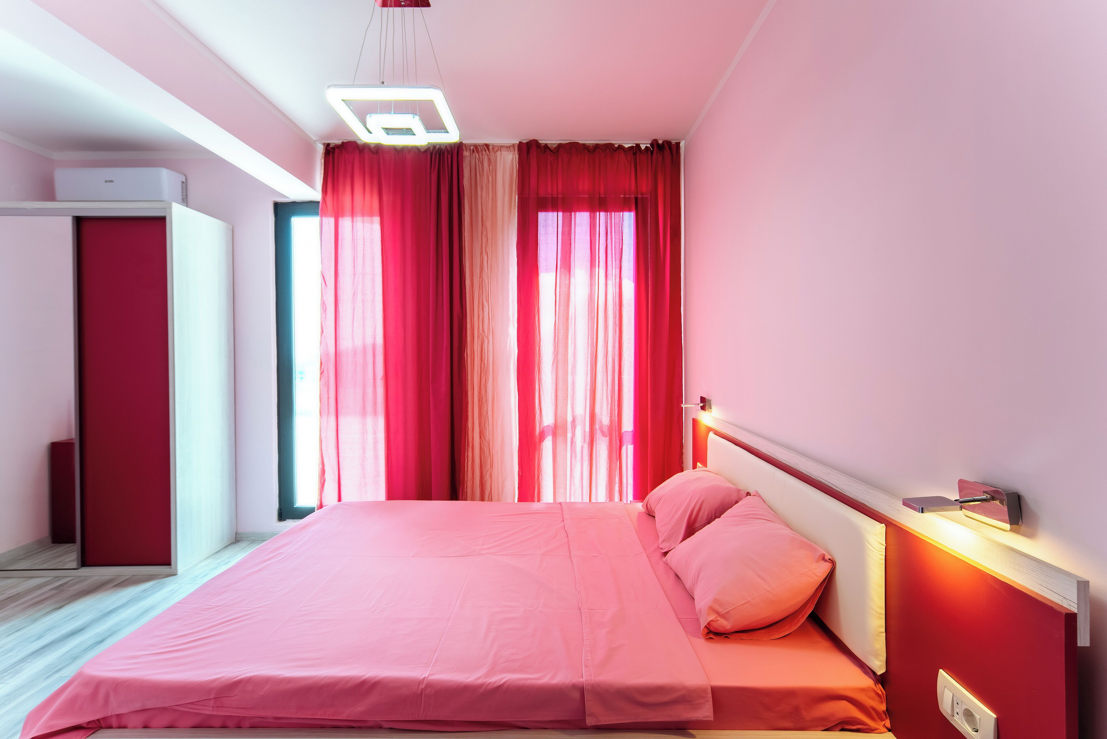 A modern bedroom with a pink bed, a wardrobe, and a window with red curtains.
