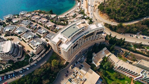 An aerial view of a coastal hotel with a curved building, parking areas, and a road leading to the beach. - Harmonia Colours (Budva)
