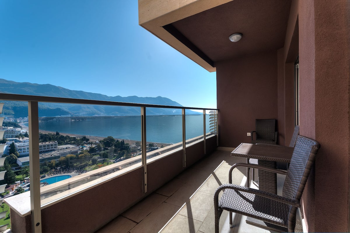 Un balcón con vista a un lago y montañas, una mesa y sillas de mimbre.