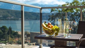 A balcony with a glass railing, a wicker table, a bowl of fruit, and a bottle of wine. - Harmonia Colours (Budva)