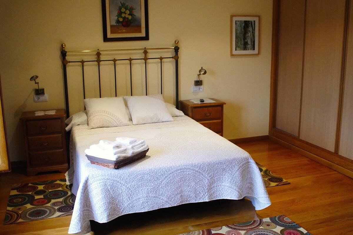 Un lit bien fait, recouvert d’une couvre-lit blanche et agrémenté d’oreillers, une table de chevet avec une lampe, une petite table en bois et un tapis à motifs sur le sol.