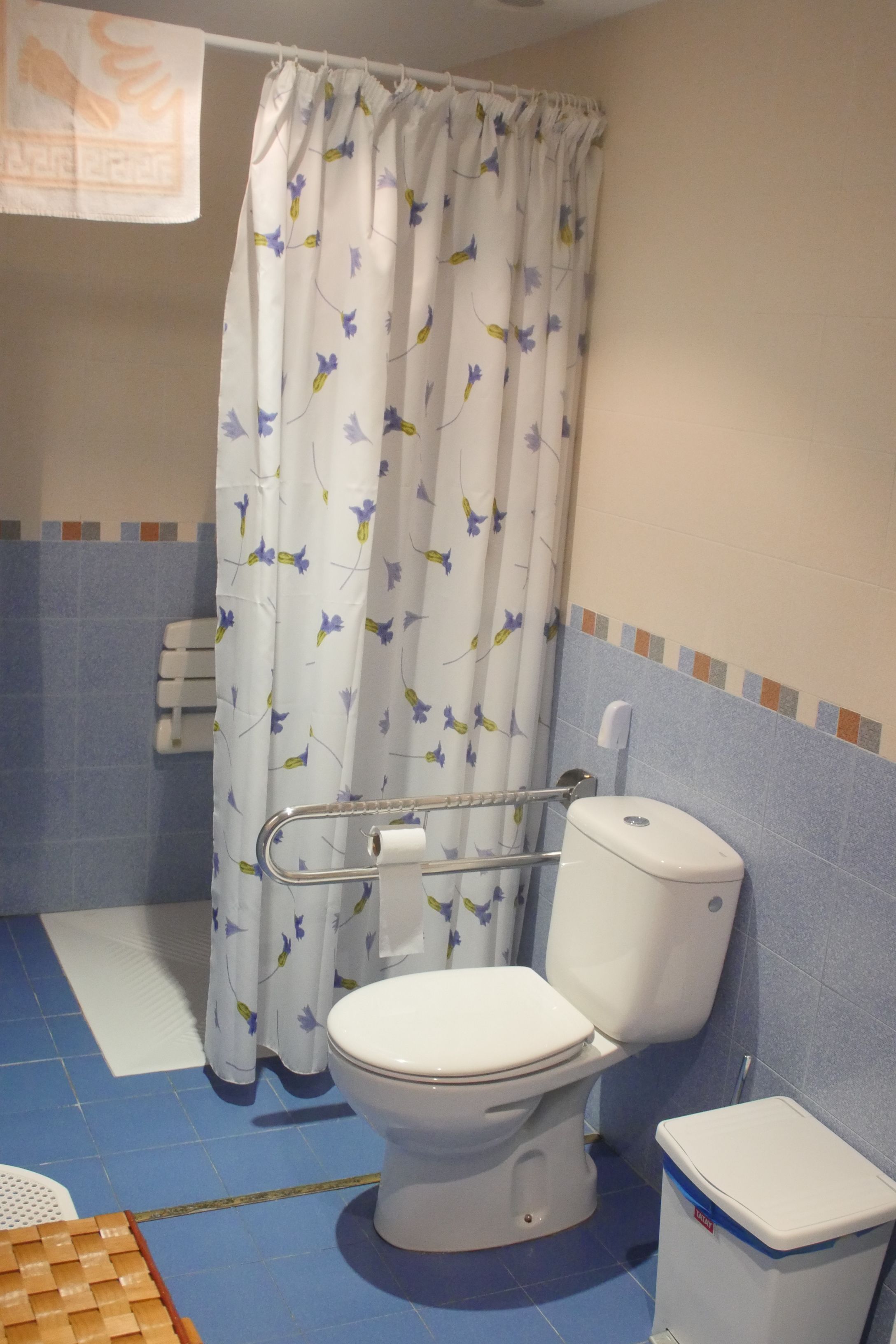 A bathroom with a toilet, a shower curtain with a floral pattern, a handrail, a waste bin, and a towel dispenser.