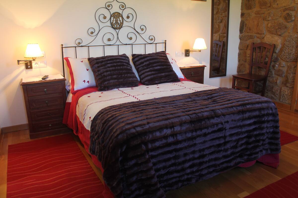 Un lit bien fait, recouvert d’une couverture sombre à rayures, deux tables de chevet avec des lampes, une table de nuit en bois et un mur en pierre.