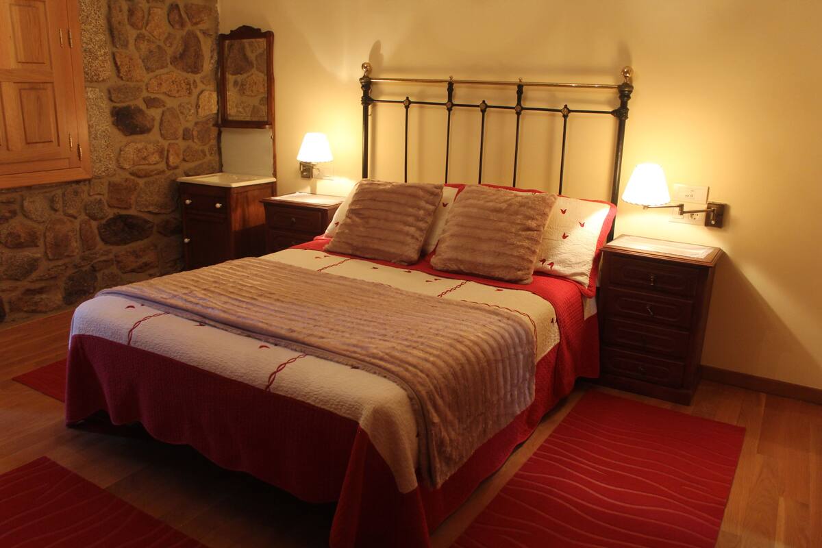 Un lit bien fait, recouvert d’une couvre-lit rouge et blanche, deux tables de chevet avec des lampes, un mur en pierre et un plancher en bois.