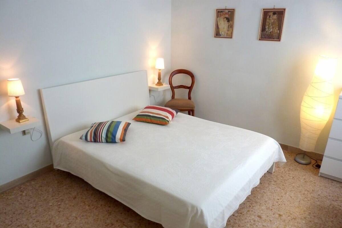 A neatly made bed with a white bedspread and striped pillows, a wooden nightstand with a lamp, and two framed artworks on the wall.