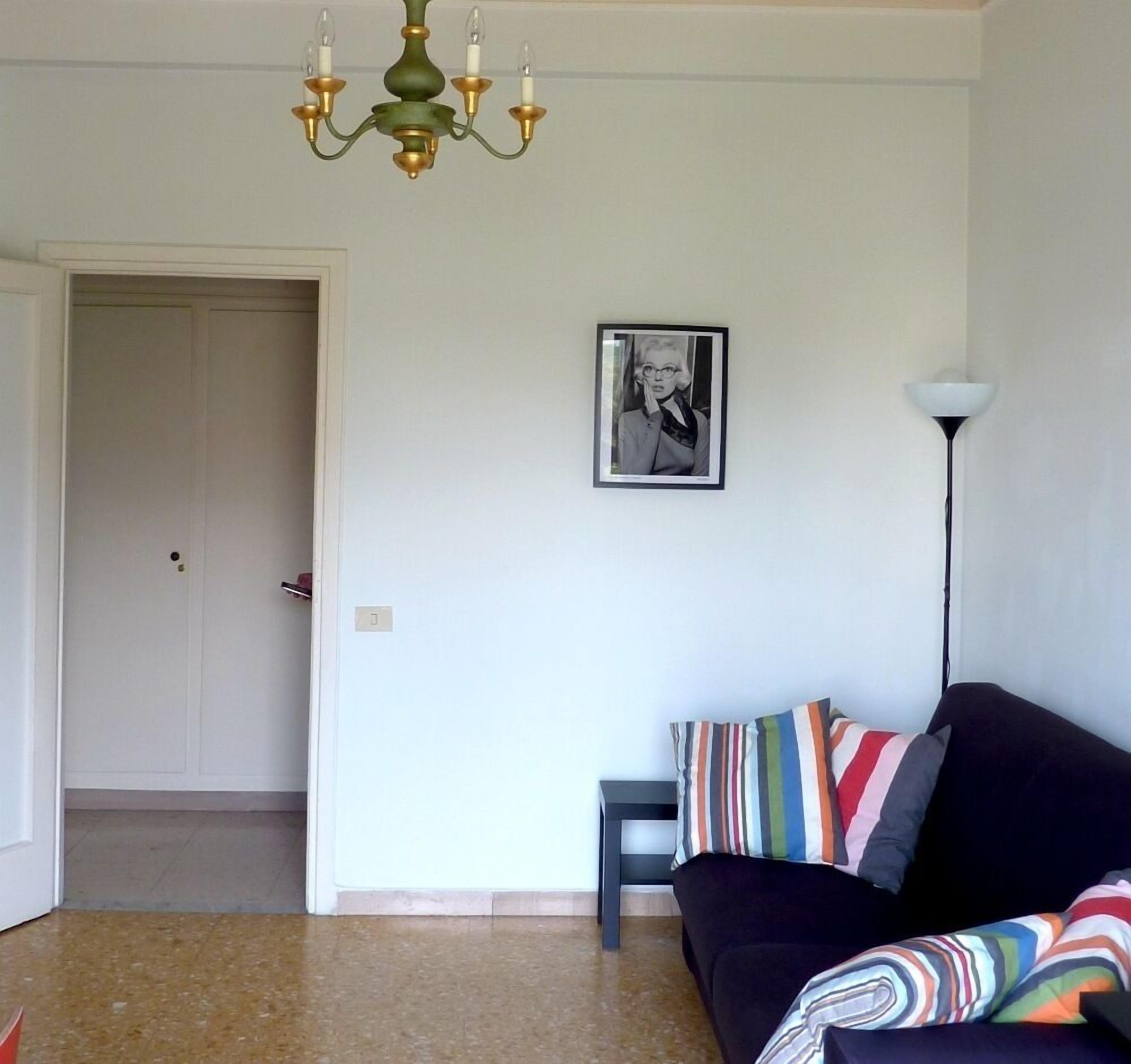 Un salon avec un canapé foncé, des coussins rayés aux couleurs vives, un tableau encadré au mur, un grand lampadaire et un lustre.